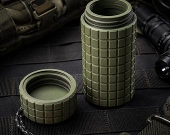 Può includere: Un contenitore cilindrico color verde oliva con un motivo a griglia testurizzato sull'esterno. Il contenitore presenta un coperchio a vite con un cordino bianco e nero attaccato a una piccola perlina di plastica rossa, con filettatura interna.