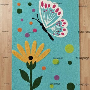 Może przedstawiać: Turkusowy dywan z fantazyjnym wzorem przedstawiającym motyla i żółty kwiat. Motyl ma białe skrzydła z różowymi akcentami, a kwiat ma żółte płatki. Kolorowe kropki są rozrzucone po dywanie.