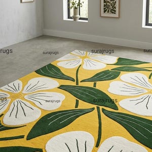 Może przedstawiać: Prostokątny dywan z kwiatowym wzorem. Dywan ma duże białe kwiaty i zielone liście na żółtym tle. Dywan znajduje się w pokoju z oprawionymi dziełami sztuki i oknem.