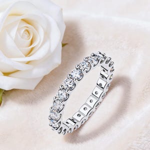 Op de afbeelding: Een zilveren eternity ring met een doorlopende rij ronde, heldere edelstenen. De ring is geplaatst tegen een zachte, witte achtergrond met een witte roos. Het ontwerp van de ring is elegant en klassiek.