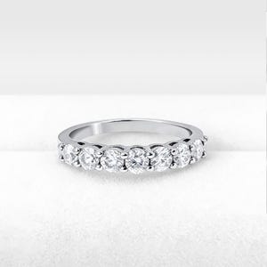 Könnte beinhalten: Ein silberner Bandring mit sieben runden, klaren Edelsteinen. Die Edelsteine sind gleichmäßig verteilt und in einer Kanalfassung gesetzt. Der Ring wird auf einer weißen Oberfläche präsentiert, die das polierte Metall und die funkelnden Steine hervorhebt.