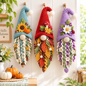 May include: Three hanging crochet gnome decorations with pointed hats. The left gnome has a blue hat with a sunflower and pumpkin. The center gnome has a red hat with mushrooms and autumn leaves. The right gnome has a purple hat with daisies.