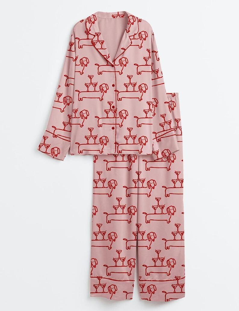 May include: Pale pink pyjama set featuring a repeating pattern of red dachshunds and champagne glasses. The long-sleeved top has a button-down front and a collar. The trousers have a comfortable, relaxed fit. Made from a soft, lightweight fabric.