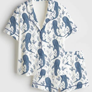 May include: White pyjama set with a blue whale shark pattern. The short-sleeved top and shorts feature a button-down design with blue trim. The whale shark design is repeated throughout the set.