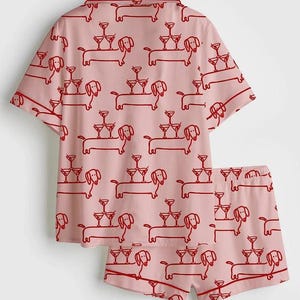 May include: A light pink pajama set with a repeating red pattern of dachshunds carrying martini glasses. The set comprises a short-sleeved shirt and matching shorts, ideal for relaxing at home.