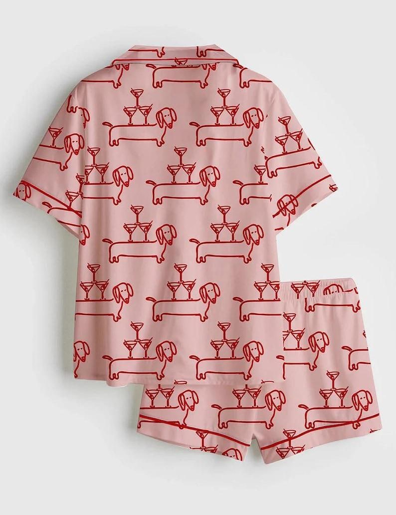 May include: A light pink pajama set with a repeating red pattern of dachshunds carrying martini glasses. The set comprises a short-sleeved shirt and matching shorts, ideal for relaxing at home.