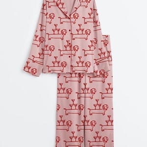 May include: A pink pyjama set featuring a repeating pattern of dachshunds balancing champagne glasses. The long-sleeved top has a button-down front and a notched collar. The trousers are straight-legged. The print is in a darker red.