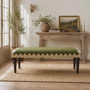 Puede incluir: Un banco rectangular con una parte superior de tela tejida en color verde y crema con un patrón de zigzag repetitivo en los bordes. El banco se apoya sobre cuatro patas de madera torneada de color marrón oscuro como base.