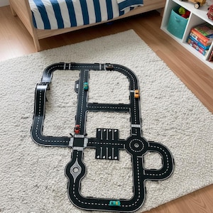 May include: A black and white toy road play mat with a variety of road features, including intersections, parking spaces, and a roundabout. Several toy cars are placed on the road. The mat is on a white rug near a bed and a shelf.