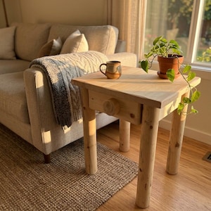 May include: A light-colored wooden side table with a single drawer featuring a round knob and four cylindrical legs. A small ceramic mug and a potted green plant with trailing vines are placed on the flat square top surface of the table.
