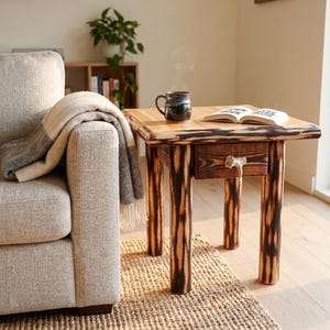 May include: A square wooden side table with a natural light wood top and four cylindrical legs featuring a charred, dark brown finish. The table includes a single drawer with a light-colored antler handle, positioned next to a sofa.