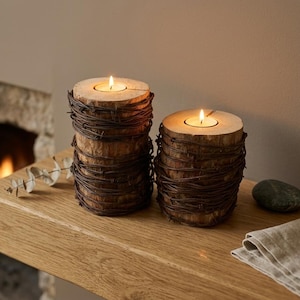 May include: Two cylindrical wooden candle holders wrapped in dark brown barbed wire. Each holder features a flat top surface with a circular cutout containing a lit tea light candle, creating a warm glow against the natural wood grain texture.