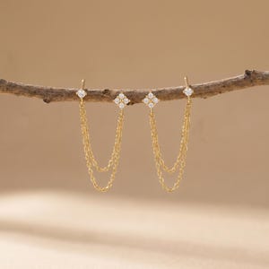 May include: Gold-coloured earrings with a delicate design. Each earring has a small, sparkling diamond at the top, connected to a chain that dangles down. The chain leads to a cluster of four diamonds. The earrings are displayed on a brown branch.