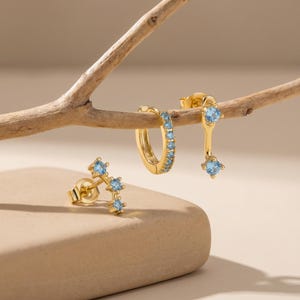 May include: Three pairs of gold-colored earrings with light blue gemstones. One pair is a stud design with three stones, another is a hoop style with multiple stones, and the third is a drop earring with two stones.
