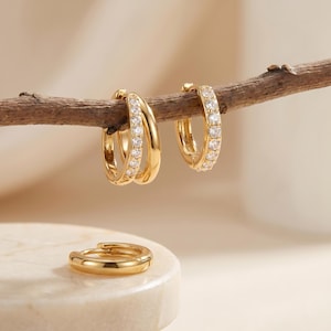 May include: Gold-colored hoop earrings with clear, sparkling stones. One pair features a double hoop design, while the other is a single hoop. A smaller, single gold hoop earring rests on a white surface. The earrings are displayed on a brown branch.