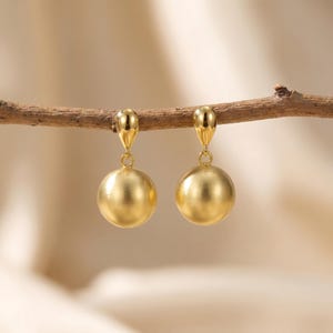 Puede incluir: Un par de pendientes colgantes de tono dorado con un colgante esférico de acabado mate liso suspendido de un componente de perno en forma de gota pulida. Los pendientes se muestran colgados de una rama de madera fina y horizontal.