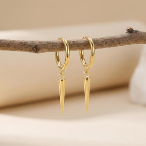 May include: Two gold-coloured hoop earrings, each with a pointed, cone-shaped charm suspended from the hoop. The earrings are presented on a brown branch against a soft, neutral backdrop. The earrings have a polished, shiny finish.