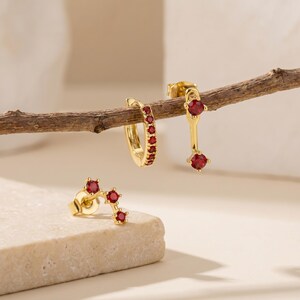 Könnte beinhalten: Drei Paar goldfarbene Ohrringe mit roten Edelsteinen. Ein Paar ist ein Stecker-Design, ein anderes ist ein Creolen-Stil, und das dritte ist ein Hängeohrring. Die Ohrringe werden auf einer hellen Oberfläche und einem Ast aus Holz präsentiert.