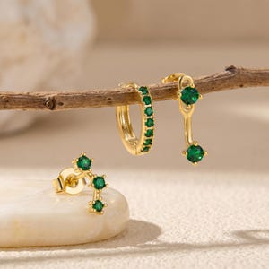 May include: Three pieces of gold jewellery: a pair of stud earrings with three green gems, a hoop earring with green gems, and a drop earring with two green gems. The jewellery is displayed on a wooden branch and a white stone.