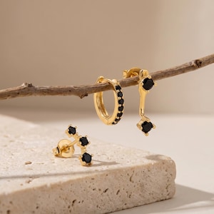 May include: A collection of gold-colored earrings with black gemstones. The earrings include a pair of small hoop earrings, a pair of drop earrings, and a pair of stud earrings. The earrings are displayed on a neutral background.