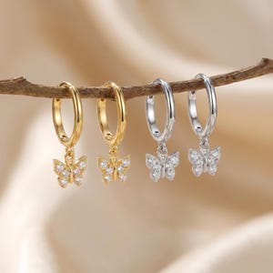May include: Two pairs of hoop earrings with butterfly charms. One pair is gold-toned, the other is silver-toned. Each butterfly charm is encrusted with small, clear gemstones. The earrings are hanging from a brown twig against a soft, neutral background.