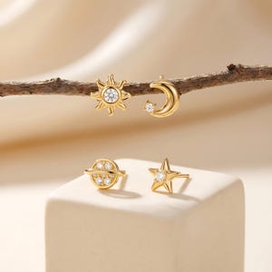 May include: Gold-colored celestial-themed stud earrings. The set includes a sun, moon, planet, and star design, each with a central clear gemstone. The earrings are displayed on a neutral background, showcasing their intricate details.