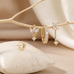 Könnte beinhalten: Eine Sammlung goldfarbener Ohrringe, darunter ein Halbmond-Ohrstecker, ein floraler Ohrhänger, ein Creolen-Ohrring und ein blütenförmiger Ohrhänger, alle mit klaren, funkelnden Edelsteinen verziert. Die Ohrringe werden auf einer hellen Oberfläche präsentiert.