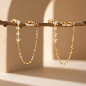 May include: A pair of gold-colored ear cuffs with dangling chains and sparkling clear gemstones. The ear cuffs are connected by a delicate chain, and the design features a stud earring and a hoop earring.