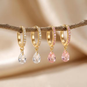 Könnte beinhalten: Vier goldfarbene Creolen mit tropfenförmigen Edelsteinen. Zwei Ohrringe haben klare Steine und zwei rosa Steine. Die Reifen sind mit kleinen, klaren oder rosa Steinen verziert.