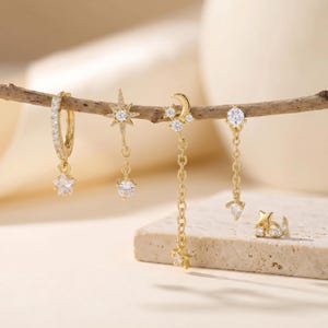 May include: A collection of gold-toned earrings featuring celestial designs. The earrings include a hoop with a star charm, a starburst drop, a moon and star drop, a single star drop, and a star and crescent ear cuff, all adorned with clear gemstones.