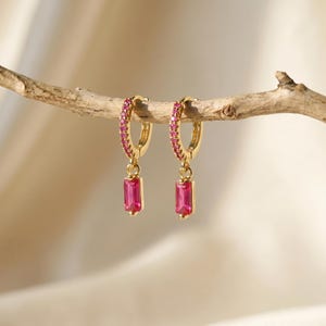 May include: A pair of gold hoop earrings featuring a row of small pink stones along the hoop and a single rectangular pink baguette-cut stone dangling from the bottom of each earring, suspended from a thin, light-coloured wooden branch.