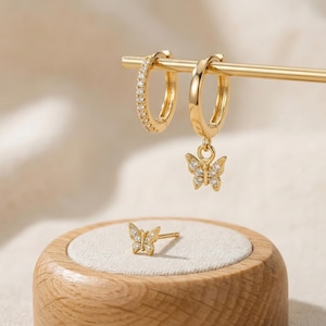 May include: Gold-toned jewelry set featuring two hoop earrings, one with a row of small clear stones and the other with a dangling butterfly charm. A matching butterfly stud earring rests on a wooden display.