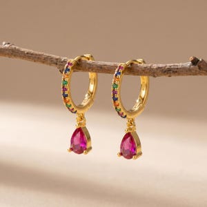 May include: A pair of gold-colored hoop earrings. Each earring features a row of colorful gemstones and a teardrop-shaped, magenta-colored pendant. The earrings are displayed on a small, brown branch against a neutral background.