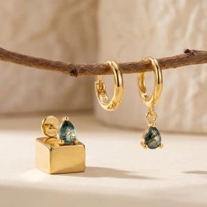 Puede incluir: Pendientes dorados, que incluyen un par de pendientes de aro y un pendiente de botón con una base cuadrada. Los pendientes presentan una piedra verde en forma de lágrima con un patrón único. Los pendientes se exhiben en una rama de madera.