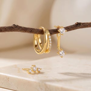 Puede incluir: Una colección de pendientes dorados. Un par es un diseño de doble aro con pequeñas piedras transparentes. Otro par presenta un pendiente con una piedra transparente cuadrada colgante. Un tercer pendiente es un pendiente con tres piedras transparentes.