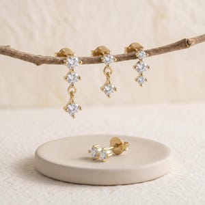 May include: Gold-colored stud earrings and dangling earrings with clear, sparkling cubic zirconia stones. The earrings are displayed on a neutral background, showcasing their delicate design and craftsmanship. The earrings are a classic jewelry item.