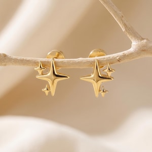 May include: A pair of gold-colored star-shaped stud earrings. Each earring features a large four-pointed star with two smaller stars. The earrings are displayed on a light-colored branch against a soft, neutral background.
