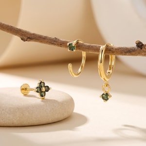 May include: Gold-toned earrings with green gemstones. The image features a stud earring with a four-leaf clover design, a small hoop earring, and a larger hoop earring with a dangling gemstone. The earrings are displayed on a wooden branch and a stone.