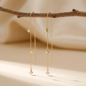 May include: Gold-colored chain earrings with multiple clear, sparkling cubic zirconia stones. The earrings are hanging from a brown branch against a soft, neutral background. The earrings have a delicate, elegant design.