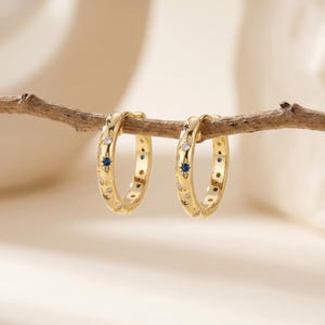 Könnte beinhalten: Ein Paar goldene Creolen, verziert mit kleinen, funkelnden weißen und blauen Edelsteinen. Die Ohrringe sind mit einem Himmelsdesign mit Sternen und Punkten verziert. Sie werden auf einem dünnen, braunen Ast vor einem weichen, neutralen Hintergrund präsentiert.