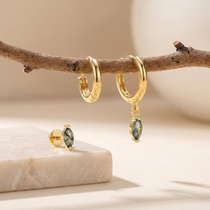 May include: Gold-colored hoop earrings and a stud earring with a green stone. One hoop earring hangs from a branch, and the other hoop earring has a green stone pendant. The stud earring is on a white surface.