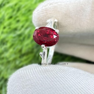 Op de afbeelding: Een zilveren ring met een grote, ovaal geslepen, dieprode edelsteen. De band van de ring heeft een delicaat, takachtig ontwerp. De ring wordt tegen een achtergrond van groen gras gehouden.