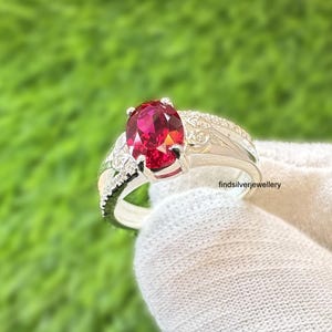Può includere: Un anello d'argento con una grande pietra preziosa rossa brillante a taglio ovale. La fascia è rifinita con pietre più piccole e trasparenti e disegni intricati. L'anello è tenuto su uno sfondo verde.