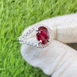 May include: A silver ring featuring a large, oval-cut, deep red gemstone, surrounded by a halo of smaller, clear stones. The ring's band splits into two, creating an open design. The ring is set against a green background.