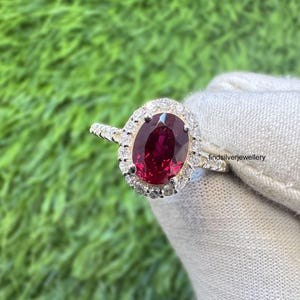 Puede incluir: Un anillo de plata con una gran gema roja oscura de forma ovalada en el centro, rodeada por un halo de piedras más pequeñas y claras. El anillo se encuentra sobre un fondo verde.