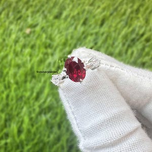 Può includere: Un anello d'argento con una grande pietra preziosa rosso scuro a taglio ovale come elemento centrale, affiancata da due pietre più piccole e trasparenti. L'anello è mostrato su un tessuto bianco, con uno sfondo verde erboso.
