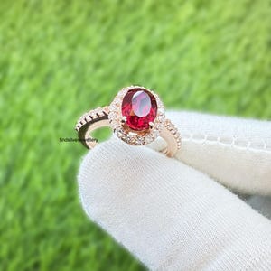 Peut inclure: Bague en or rose ornée d'une grosse pierre rouge ovale, entourée de petits brillants. La monture est également ornée de petits brillants. La bague est posée sur un fond vert.