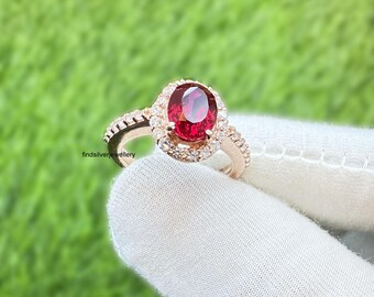 May include: Rose gold ring with a large oval-cut red gemstone, encircled by smaller clear stones. The band also features small clear stones. The ring is set against a green background.