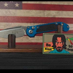 May include: A blue and black folding knife with a silver blade rests on a wooden stand. Beside it is a patch with the text "Be Kind to Dogs" and a graphic of a man. The background is an American flag.