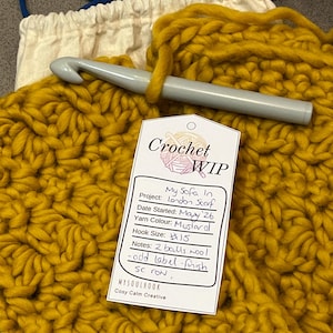 May include: A mustard yellow chunky crochet project in progress with a grey crochet hook resting on top. A white paper tag labeled Crochet WIP is attached, featuring handwritten details including Project: London Scarf, Date Started: May '26, Yarn Colour: Mustard, Hook Size: 15, and Notes: 2 balls wool - add label - finish sc row.